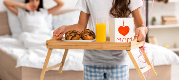 cropped view of child holding tray with breakfast, mothers day card with heart sign and mom lettering, while mother stretching in bed